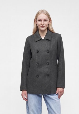 Giovane donna con lunghi capelli biondi che indossa un cappotto doppiopetto grigio scuro e jeans azzurri, in piedi davanti a uno sfondo semplice.