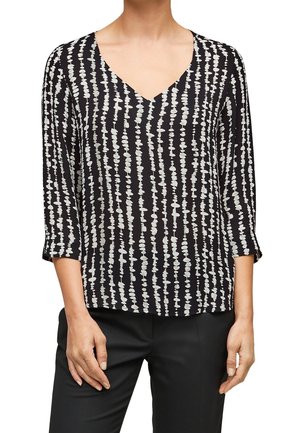 Femme portant un chemisier noir avec des rayures verticales blanches en pointillés et des manches trois-quarts, associé à un pantalon noir.
