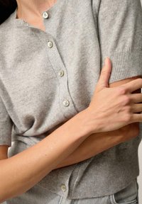 Szary kardigan z krótkim rękawem, wykonany z miękkiego materiału, posiada okrągły dekolt, cztery guziki oraz ściągacze na rękawach i u dołu.