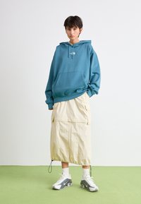 Haut à capuche teal avec une poche avant et un cordon de serrage, associé à une jupe crème arrivant au genou avec des poches et un ourlet à cordon de serrage. Baskets blanches.
