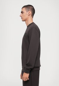 Mørkebrun hettegenser med lange ermer, rund halslinning og et subtilt teksturert merke på brystet. Enkelt, minimalistisk design.