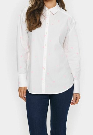 Femme portant une chemise blanche à manches longues avec de petits motifs de cœurs roses et un jean bleu foncé, debout sur un fond blanc.