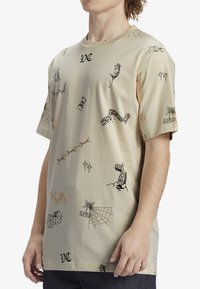 Camiseta de algodón beige con mangas cortas, que presenta varios estampados gráficos que incluyen objetos, patrones y logotipos en contraste negro y marrón.