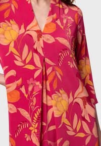 Abito floreale in un vivace rosa con fiori arancioni e gialli. Presenta un scollo a V, maniche a tre quarti e un dettaglio di piega sottile.