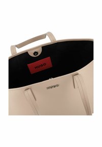 Sac fourre-tout beige avec un extérieur en synthétique texturé, deux poignées supérieures, un intérieur noir et une étiquette de marque rouge. Accents en métal argenté visibles.