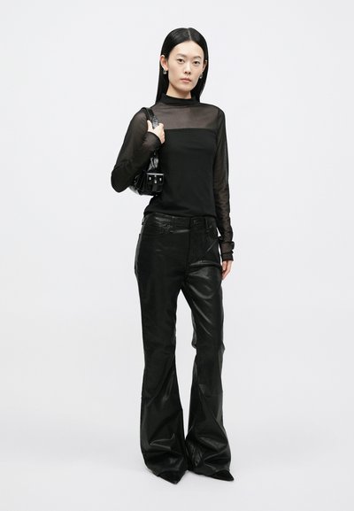 Top preto transparente de manga comprida combinado com calças pretas, brilhantes e evasê. A modelo segura uma pequena mala preta com alças e detalhes.