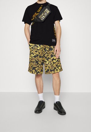 Schwarzes T-Shirt mit gelben grafischen Details, gemusterte gelb-schwarze Shorts und schwarze Sneakers mit goldenen Akzenten. Weiße Knöchelsocken.