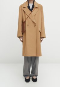 Kameliin värinen kaksirivinen takki leveillä kaula-aukolla, yhdistetty harmaisiin ruutukuosisiin housuihin, mustiin slip-on-kenkiin ja ruskeaan olkalaukkuun.