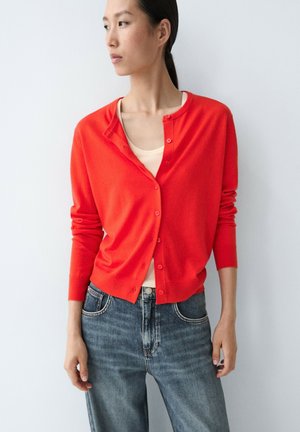 Frau trägt eine leuchtend rote, geknöpfte Strickjacke über einem hellbeigen Oberteil mit blauer Jeans, steht vor schlichtem hellem Hintergrund und schaut seitlich.