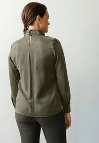 Olivengrüne Bluse mit hohem Kragen, langen Ärmeln und einem Rückenschlitz mit zwei Knöpfen. Glatte Textur, figurbetonte Silhouette.