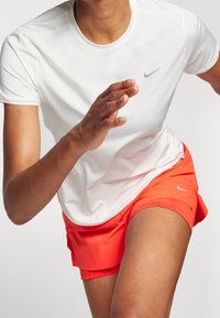Camiseta deportiva blanca de manga corta con un logo gris, acompañada de unos pantalones cortos naranja brillante que cuentan con un logo sutil y un ajuste ceñido.