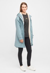 Abrigo impermeable azul claro con interior forrado de piel, cierre de cremallera y botón, capucha ajustable, combinado con jeans negros y zapatillas blancas.
