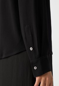Black ribbed long-sleeve shirt featuring a buttoned cuff with silver-tone buttons, soft texture, and a slightly curved hem.
