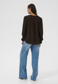 Blusa marrone a maniche lunghe con dettaglio plissettato sul retro, abbinata a jeans a gamba larga azzurri, che mostrano una vestibilità rilassata e un tessuto testurizzato.