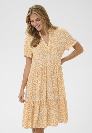 Usmievavá žena s dlhými blond vlasmi, oblečená v žltých a bielych vzorovaných šatách s krátkym rukávom, ktoré siahajú po kolená, na jednoduchom pozadí.