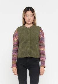 Olijfgroene fleece vest met ronde halslijn en drukknoopjes, genoten over een kleurrijk gebreid sweater met lange mouwen.