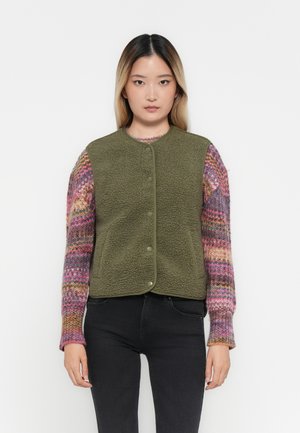 Olijfgroene fleece vest met ronde halslijn en drukknoopjes, genoten over een kleurrijk gebreid sweater met lange mouwen.