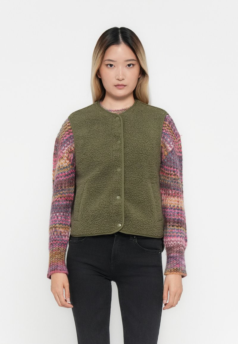 Olijfgroene fleece vest met ronde halslijn en drukknoopjes, genoten over een kleurrijk gebreid sweater met lange mouwen.