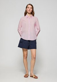 Camicia button-up rosa chiaro con colletto e logo, abbinata a pantaloni cargo blu navy. L'insieme include sandali marroni con un accento in tessuto intrecciato.