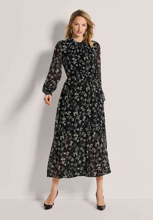 Frau trägt ein schwarzes langärmeliges Midikleid mit weißen Blumenmustern und schwarzen Absätzen, steht vor einem schlichten hellen Hintergrund.