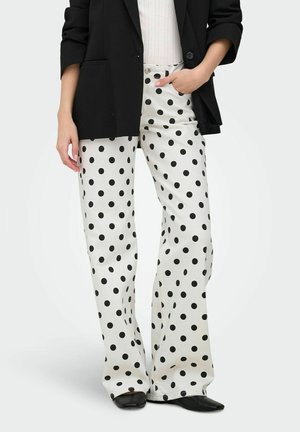 Femme portant un pantalon blanc à jambes larges avec des pois noirs, un blazer noir, un haut blanc et des chaussures noires à bout pointu, main dans la poche.