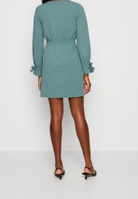 Lyhythihainen petrolinvärinen mekko, jossa on vyötäröltä kerätty muotoilu ja röyhelökäänteet. Kangas on sileää ja siinä on puhdas, minimalistinen muotoilu.