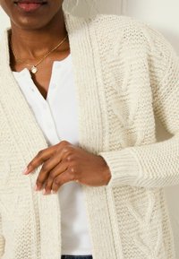 Beiger, grob gestrickter Cardigan mit Kabelmuster, offene Vorderseite, gerippte Bündchen, über einem weißen Henley-Oberteil getragen. Hand liegt auf der Vorderseite des Cardigans.