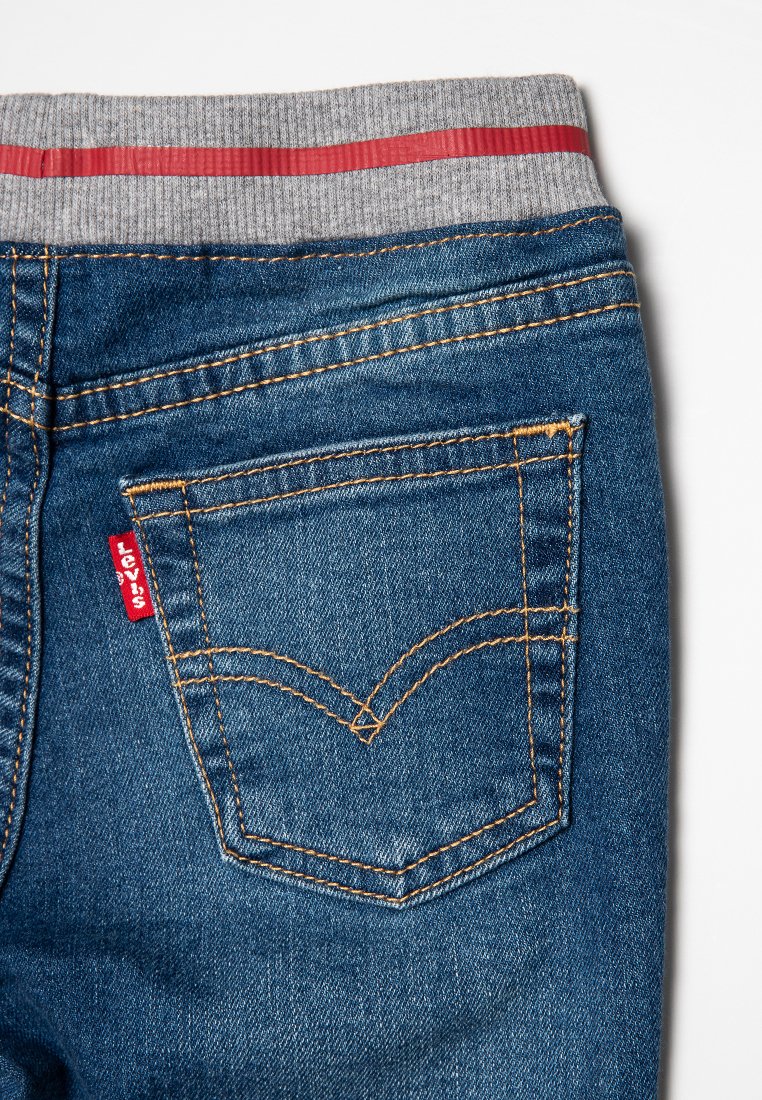Jeans de mezclilla azul con una cinturilla elástica gris que presenta una franja roja. El bolsillo trasero tiene un diseño de costura amarilla y una etiqueta roja de Levi's.