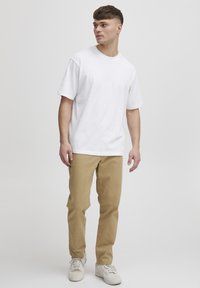 T-shirt en coton blanc à manches courtes, associé à un pantalon beige. Le mannequin se tient contre un fond uni, portant des baskets blanches.