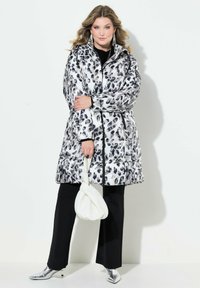 Abrigo tipo puff con un estampado de leopardo en blanco y negro, con un cuello alto, cierre de cremallera y un acabado brillante. Combinado con pantalones negros y tacones plateados.
