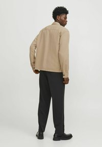 Šviesiai rusvos tekstūros marškiniai su laisvu siluetu ir mygtukais ant rankovių, derinami su juodomis kelnėmis ir juodais batais, matomi iš nugaros.