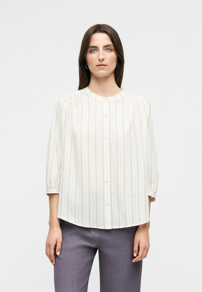 Femme portant un chemisier blanc à manches longues à boutons avec de fines rayures verticales et un pantalon gris, se tenant devant un fond uni.