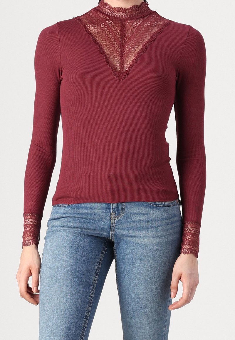 Långärmad bordeauxfärgad topp med ribbat mönster, spetsdetaljer vid halsringningen och ärmsluten, samt en figurnära silhuett. Matchad med blå jeans.
