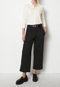 Femme portant une chemise blanche à manches longues avec des boutons, un pantalon cropped à jambes larges noirs, une ceinture noire avec une boucle dorée, et des mocassins noirs.