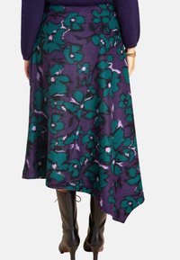 Ein lila Blumen-Wickelrock mit großen türkisfarbenen Blumen und einem asymmetrischen Saum. Getragen mit enganliegenden schwarzen knielangen Stiefeln.