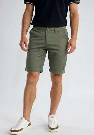 Homme portant un short olive à ourlet roulé, des baskets blanches et un t-shirt noir à manches courtes, debout devant un fond uni.