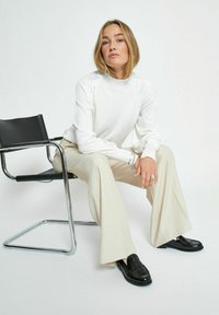Bílý top s dlouhým rukávem a nařasenými detaily na ramenou, spárovaný s béžovými širokými kalhotami a černými mokasínami, sedící na černé židli.