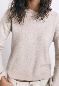 Osoba oblečená v svetlom béžovom pletenom svetri s vyhrnutými rukávmi a svetlých nohaviciach, s tmavými kučeravými vlasmi padajúcimi na plecia.