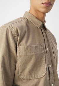 Camicia in corduroy marrone con colletto button-down, righe verticali, due tasche frontali e bottoni color crema. Texture liscia.