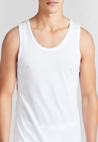 Torso maschile che indossa una semplice canottiera bianca senza maniche su uno sfondo grigio chiaro.