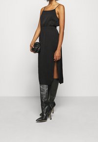 Robe noire à bretelles spaghetti, taille cintrée, fente sur le côté et coupe décontractée. Associée à des bottes en cuir noir jusqu'aux genoux et à un petit sac noir.