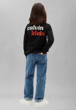 Dziecko z włosami do ramion stoi tyłem, ubrane w czarną bluzę z napisem „calvin klein”, niebieskie dżinsy i białe trampki.