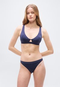 Νεαρή γυναίκα με μακριά ξανθά μαλλιά φοράει σκούρο μπλε μπικίνι με γκλίτερ και άνοιγμα σε σχήμα κλειδαρότρυπας στο πάνω μέρος, στέκεται μπροστά σε απλό ανοιχτό φόντο.