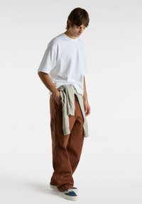 Hvid kortærmet t-shirt, olivengrønt sweatshirt bundet om livet, brune brede bukser og navy sneakers med lys accent. Simpelt, afslappet design.