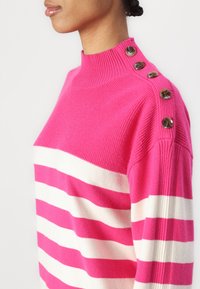 Pull en tricot avec des rayures horizontales rose vif et blanches, col haut côtelé et boutons décoratifs sur l'épaule.