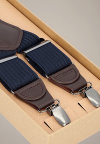 Granatowe elastyczne szelki z brązowymi skórzanymi akcentami i metalowymi klipsami, prezentowane w kartonowym pudełku. Widoczny teksturowany wzór pasków.