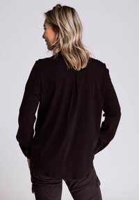 Schwarzes langärmliges Oberteil mit lockerem Schnitt und Rückenfalte, gefertigt aus weichem Stoff. Kombiniert mit dunkler, gemusterter Hose.