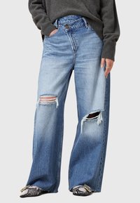 Ljusblå vida jeanstroppar med fransade, rivna knän och hög midja, tillverkade av denim. Styland med en mörk tröja och mönstrade skor.