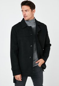 Cappotto in misto lana nero con chiusura a bottoni, design doppiopetto e spalline. Abbinato a un maglione grigio a collo alto a coste sotto.