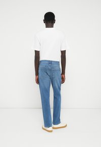 Άνδρας που φοράει λευκό μπλουζάκι, μπλε τζιν και λευκά αθλητικά παπούτσια, στέκεται με την πλάτη σε μια απλή λευκή τοίχο.